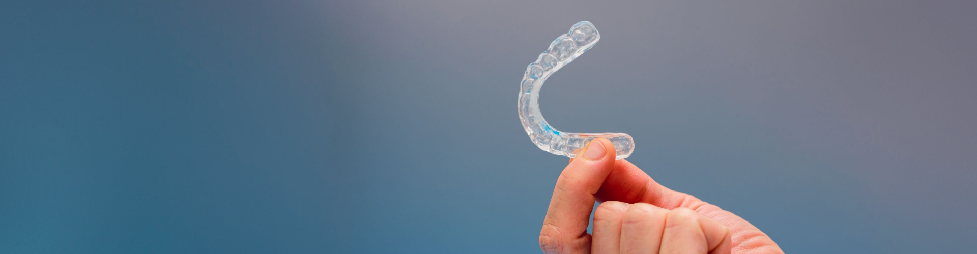 Clear aligner held between fingers against a blue background.